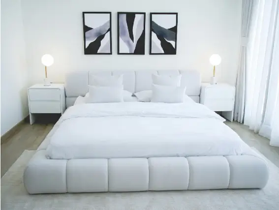 Turnkey bedroom furniture package by TUKA Dubai featuring a low-profile white tufted platform bed, matching nightstands, globe lamps, and abstract triptych wall art.